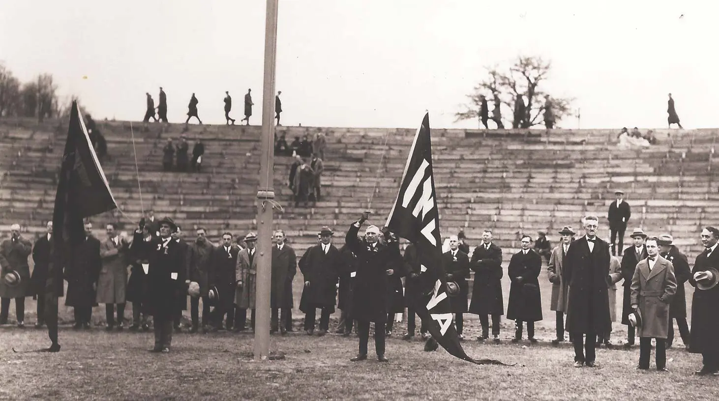 Two men in coats hoisting flags with a row of men in coats standing behind them